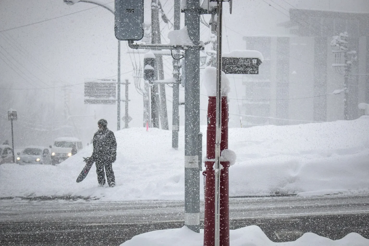 Heavy snow falling on a street in Hirafu, with a traffic signal, snow banks and a person carrying a snowboard