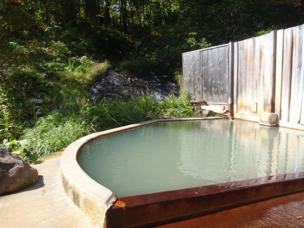 A curved open-air hot-spring bath with pale blue-green water, a wooden fence and green plants, at an inn in the Niseko Konbu Onsen area
