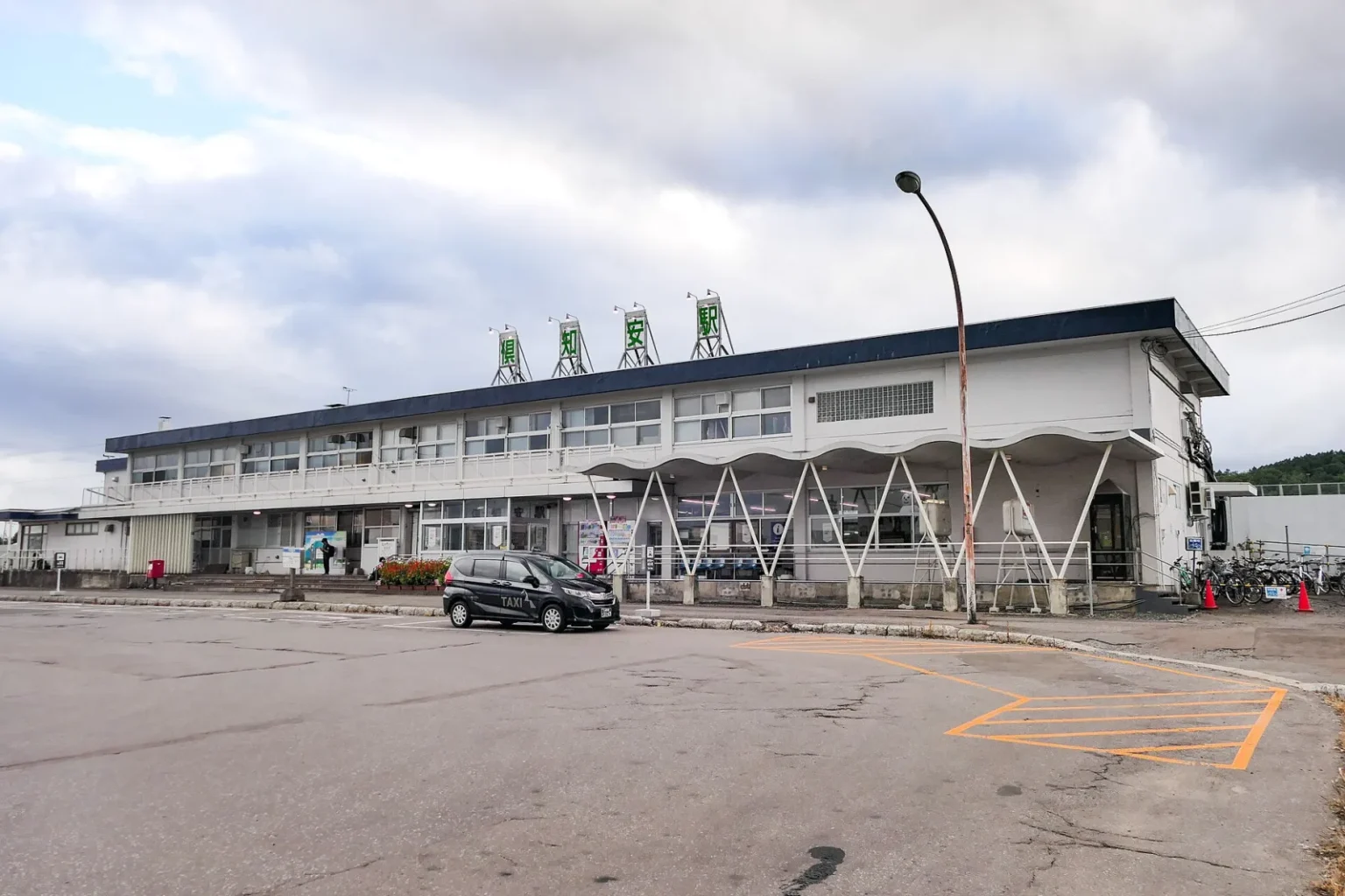 The low white building of JR Kutchan Station with its parking area in front, under a cloudy sky