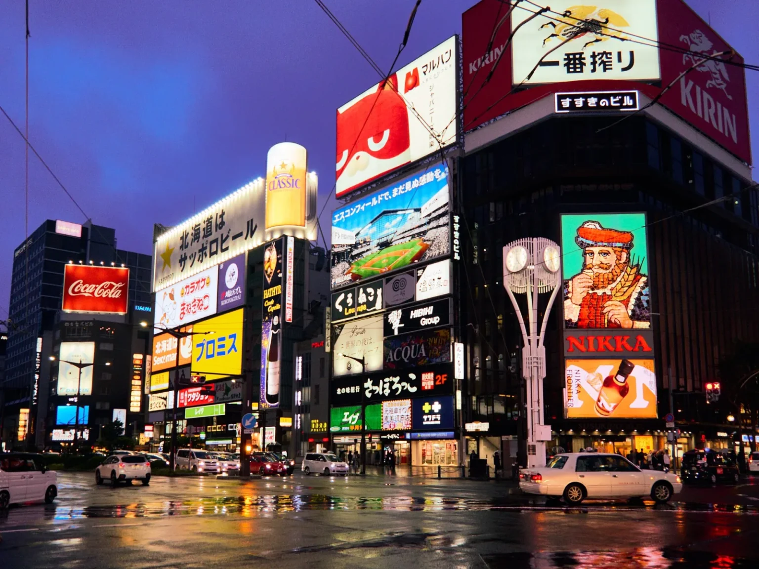 Neon signs on the buildings at the Susukino crossing in Sapporo at dusk, including the Nikka Whisky sign, with a wet road and taxis