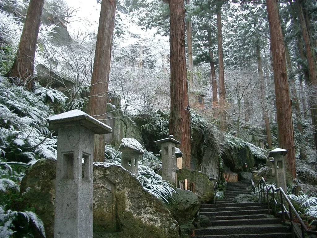 Stone lanterns and tall cedar trees dusted with snow beside a flight of stone steps with a handrail on the approach to Risshakuji
