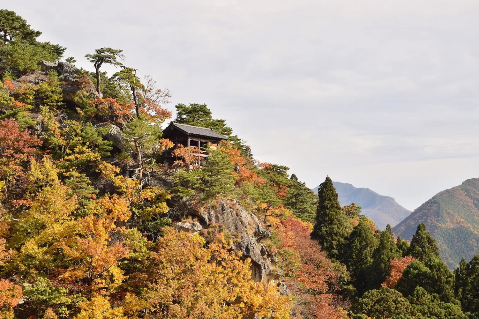 A small wooden hall on top of a rocky crag at Yamadera, surrounded by red, orange and yellow autumn trees and pines, with distant mountains under a pale sky