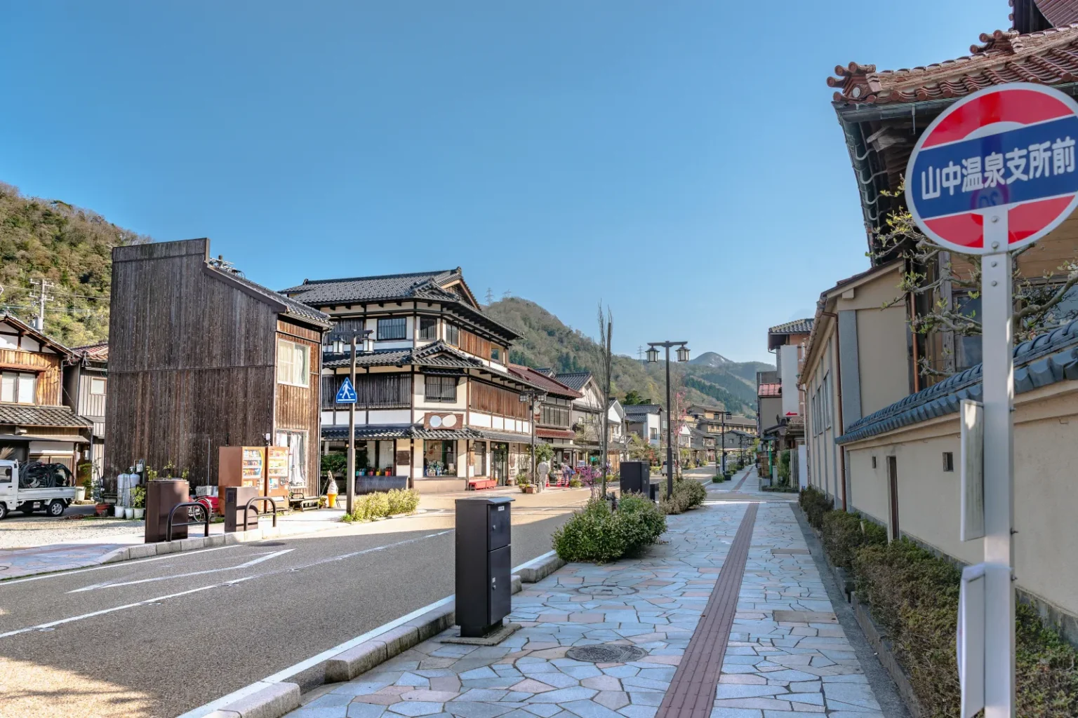 A wide street of wooden buildings with a stone-paved pavement and a bus-stop sign under blue sky