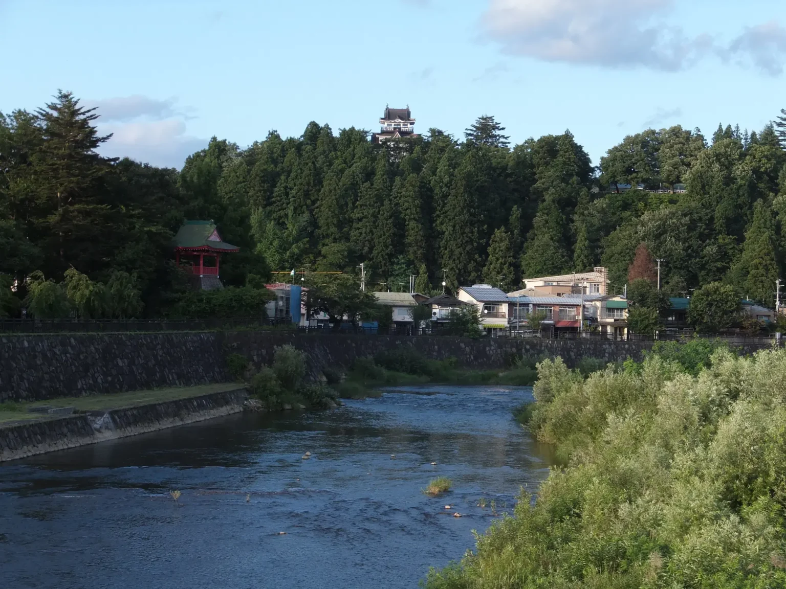 The Yokote River flowing between a stone embankment and green riverside scrub, with low houses along the far bank, a red-painted pavilion with a green roof on the left and the white Yokote Castle observatory standing above a wooded hill under a blue summer sky.