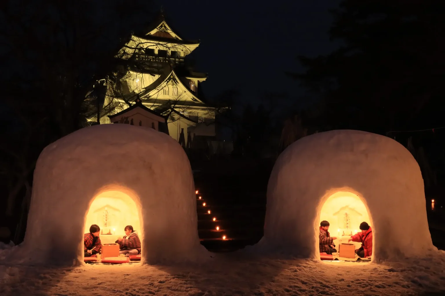 Two large dome-shaped snow huts glowing orange from candlelight at dusk, each with two children in red jackets sitting inside before a small altar, and the white keep-style observatory of Yokote Castle lit up on the hill behind.