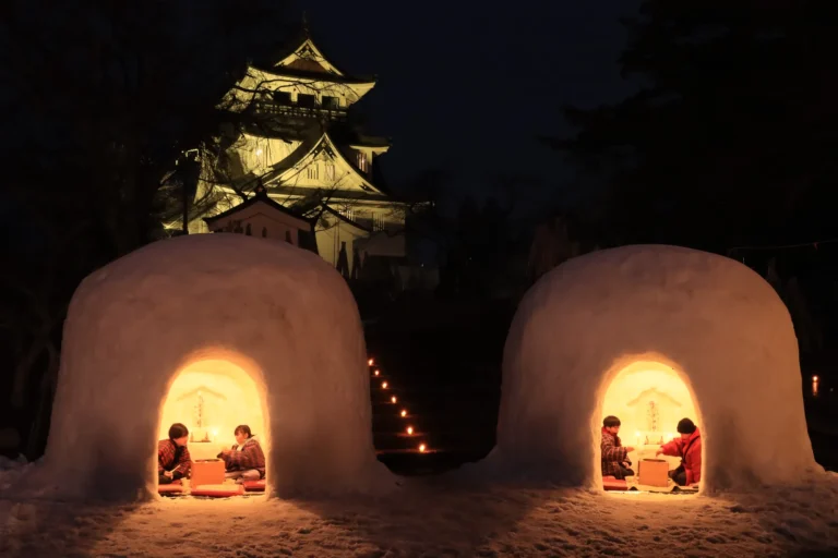 Two large dome-shaped snow huts glowing orange from candlelight at dusk, each with two children in red jackets sitting inside before a small altar, and the white keep-style observatory of Yokote Castle lit up on the hill behind.