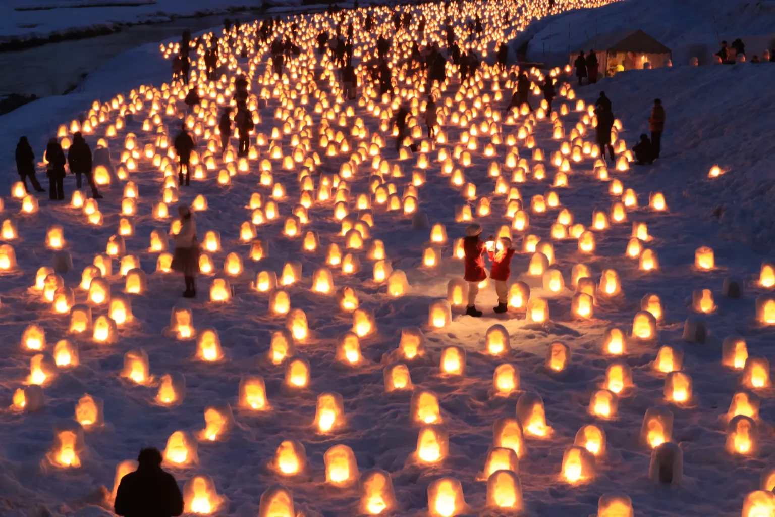 Rows upon rows of small candlelit snow domes covering a snowy riverbank at dusk, with visitors in dark coats walking between them and two children in red holding a lantern in the middle.