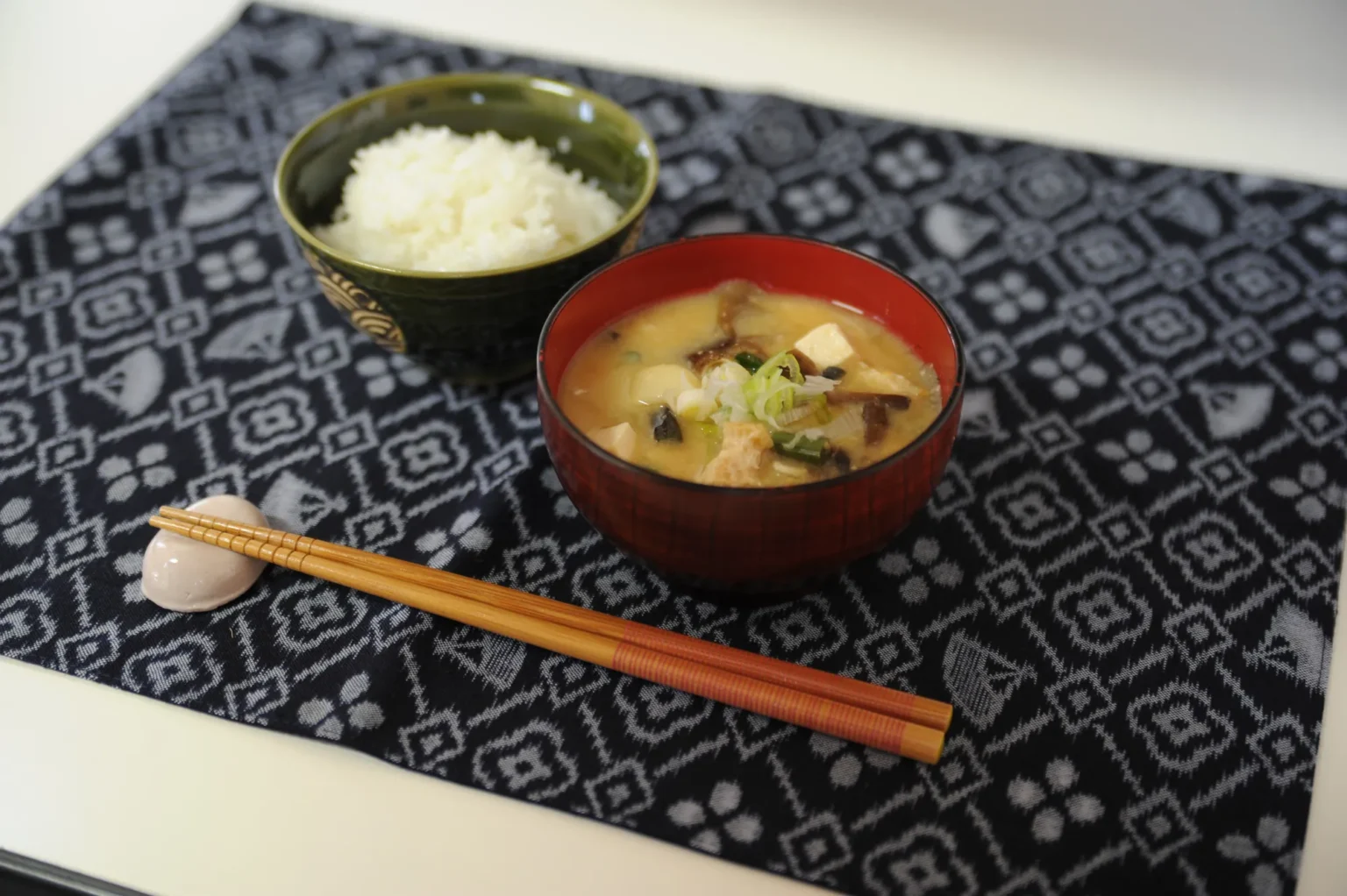 A red lacquered bowl of cloudy natto-jiru with cubes of tofu, pieces of fried tofu, dark mushrooms and sliced spring onion, beside a green bowl of white rice and bamboo chopsticks on a navy and white patterned cloth.