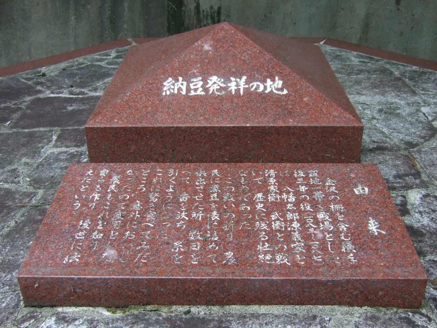 Close-up of the red granite monument: the pyramid top carved 納豆発祥の地 in white-filled characters, and in front a flat slab headed 由来, origin, with eighteen short vertical lines of carved Japanese text telling the Later Three Years' War natto story.
