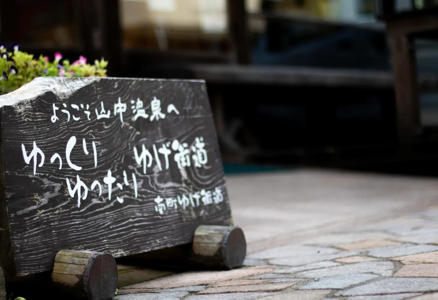 A dark wooden board with white Japanese handwriting welcoming visitors, on a stone-paved street