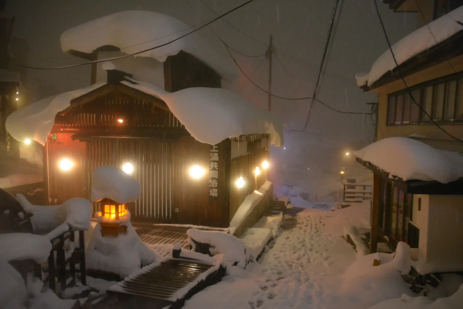 A small wooden bathhouse with deep snow piled on its roof and lamps lit along its walls, a vertical sign reading 上湯共同浴場, a snow-capped stone lantern in front and a snowy lane with footprints leading uphill at night
